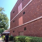 A three-story red brick building features several large Doors to Nowhere built into the upper walls, with no balconies or stairs. People stroll on the sidewalk nearby, while shrubs line the base of the building.