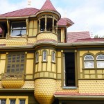A yellow and brown Victorian-style building with red shingled roofs, scalloped siding, arched windows, and a small tower features one of its famed Doors to Nowhere, marked by an open exterior door that leads outside.
