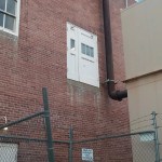 A white double door, known as "Doors to Nowhere," is installed on the second story of a red brick building with no balcony or stairs, above a concrete ledge. A large pipe runs beside it, with a chain-link fence and warning sign in the foreground.