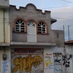 A gray two-story building with arched windows and decorative brick trim features a shuttered storefront, "Polleria Mary," graffiti on the metal door and wall, and upper-level doors to nowhere that add a curious touch to its facade.