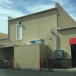 A beige commercial building features a classic "doors to nowhere" setup, with a high, inaccessible door on the upper wall. Nearby are ventilation pipes, utility boxes, and two red bollards under a partly cloudy sky.