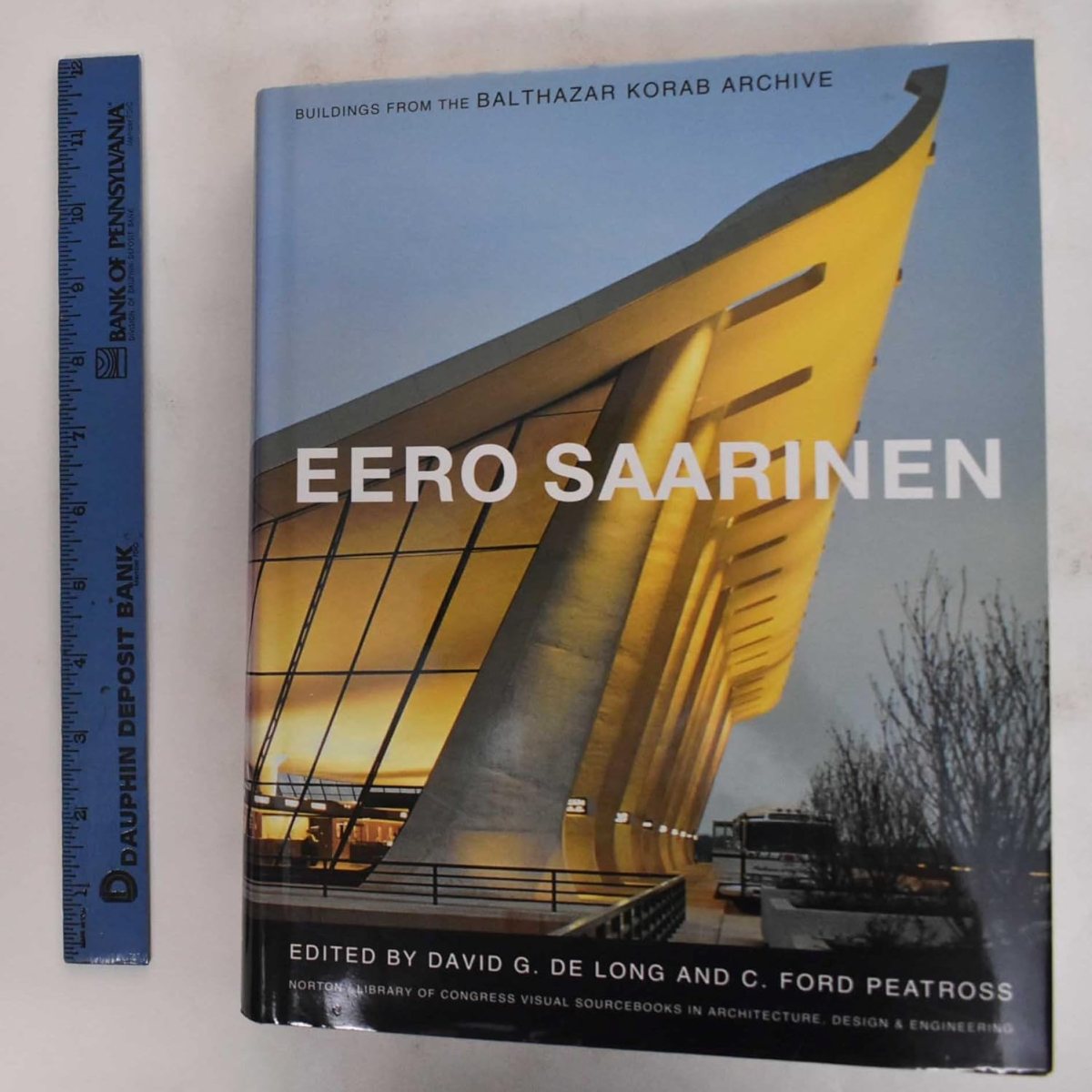 A hardcover book titled Eero Saarinen featuring a photo of a modern building with curved yellow-lit architecture, next to a blue ruler marked Bank of Pennsylvania. Ideal for any student or architect, the book is edited by David G. De Long and C. Ford Peatross.